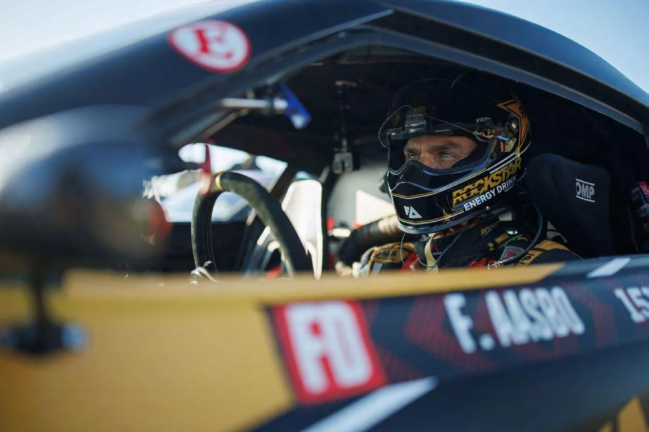 Helmeted race car driver strapped into the cockpit of a Papadakis Racing competition car.