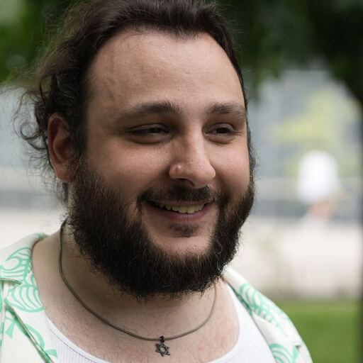 Close-up of a smiling man with a beard and long hair, wearing a white shirt and a necklace with a star pendant, outdoors with blurred green trees in the background.