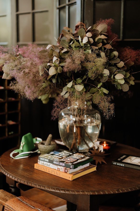 A glass vase filled with eucalyptus and dried floral branches sits on a wooden table, with a stack of books, a green camera, a candle, and a small bowl nearby.