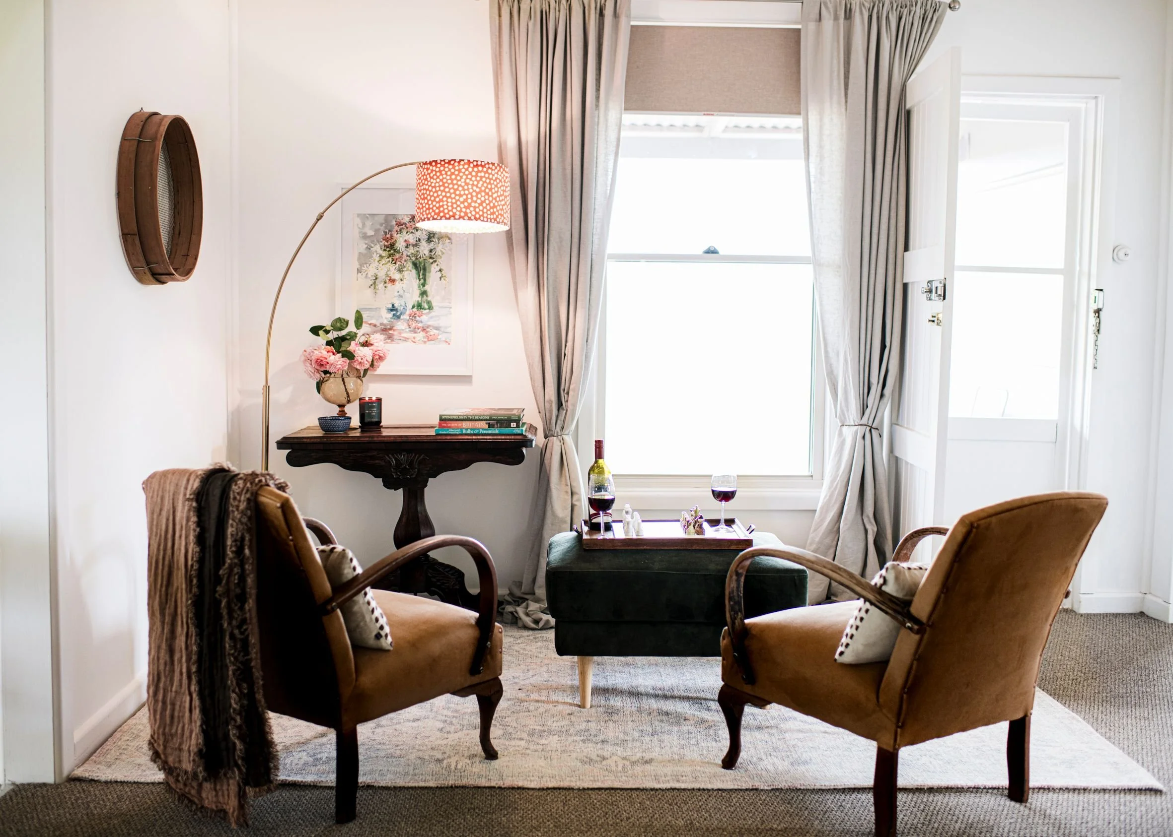 A cozy living room with four vintage armchairs arranged around a small coffee table with two glasses of red wine. There is a large window with beige curtains, a console table with a vase of pink flowers, books, and a decorative lamp, and framed artwork on the wall.