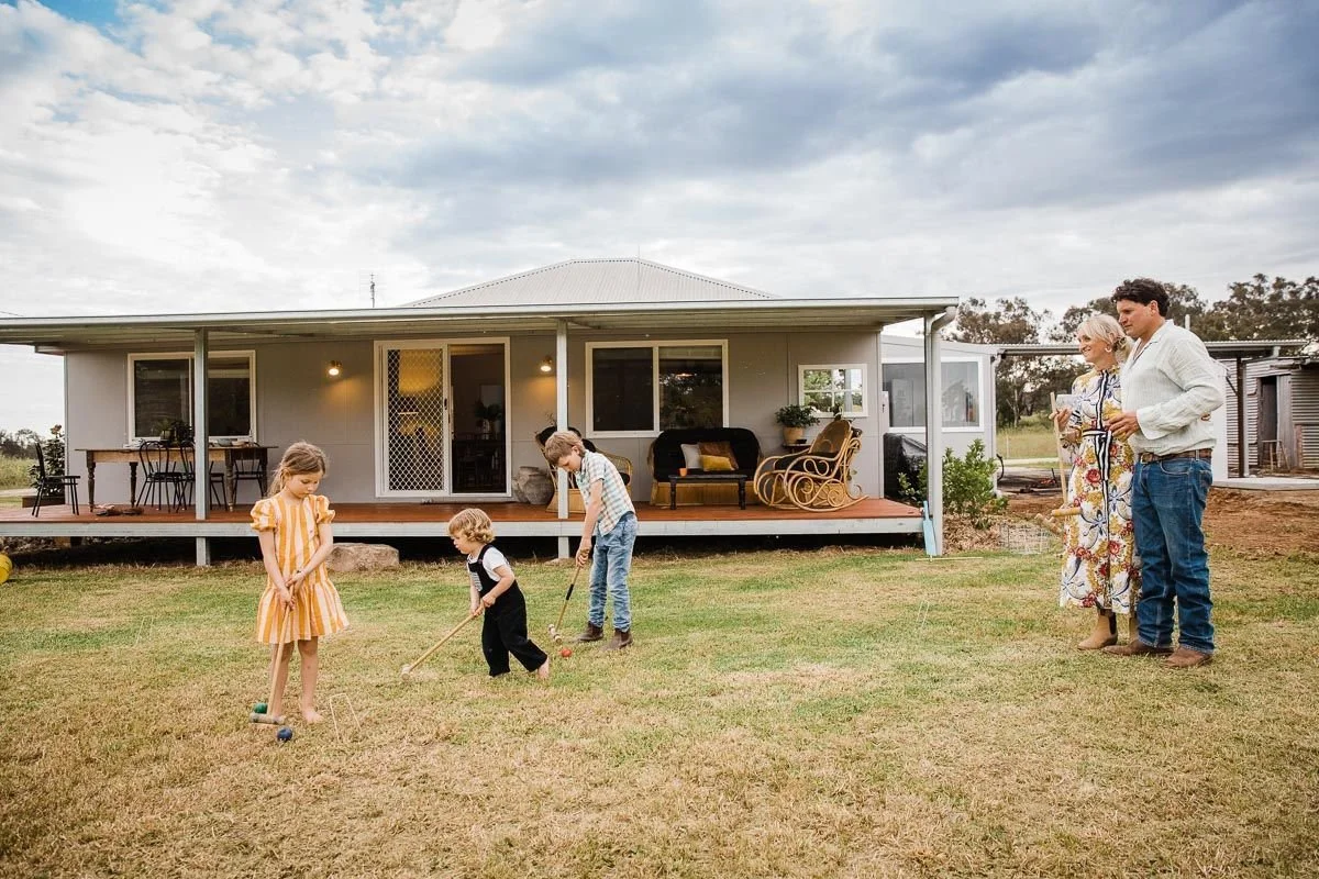 Family playing mini golf on a lawn in front of a house with porch and outdoor furniture, while two adults watch.
