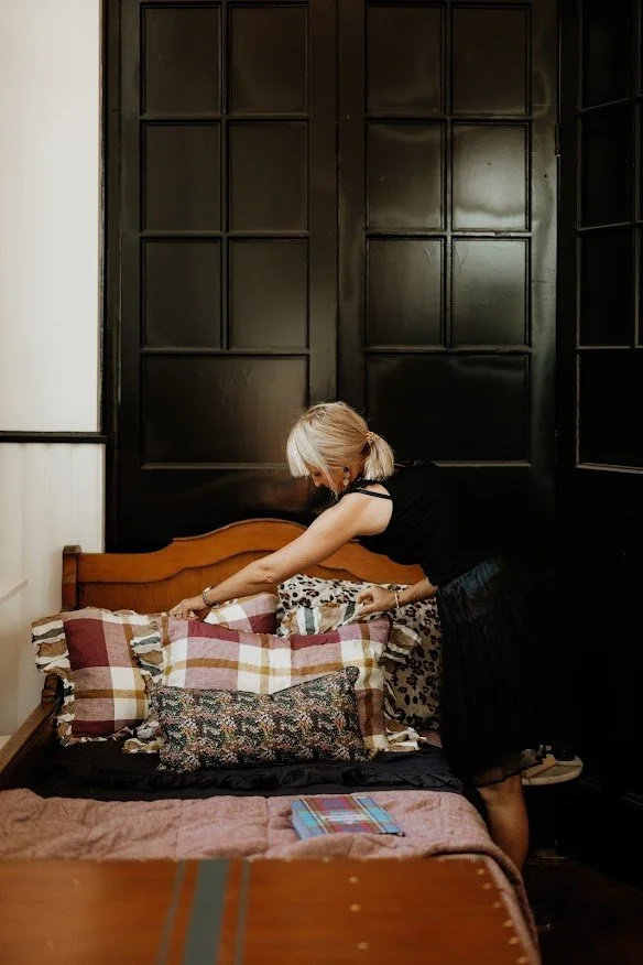 A woman with blonde hair tied in a ponytail, dressed in black, makes the bed in a bedroom. She is adjusting pillows on a wooden bed frame that has a checkered pillow, a floral pillow, and others. The room has dark paneled walls behind the bed and a light-colored wall to the side. There is a tablet or book on the bed.
