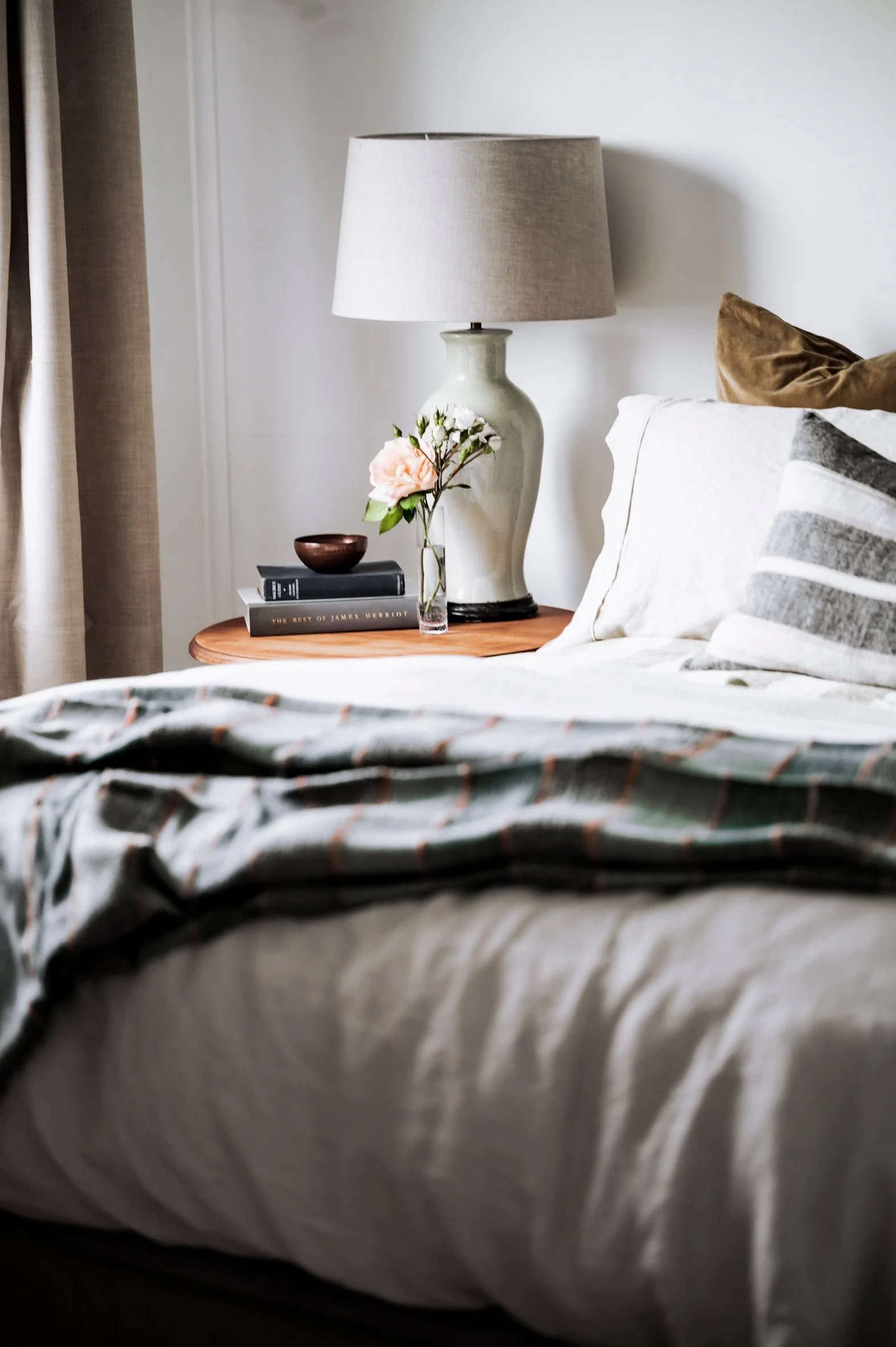 Close-up of a bedroom corner with a wooden nightstand holding a ceramic table lamp, a vase with flowers, and stacked books, next to a bed with white and striped pillows and a plaid blanket.