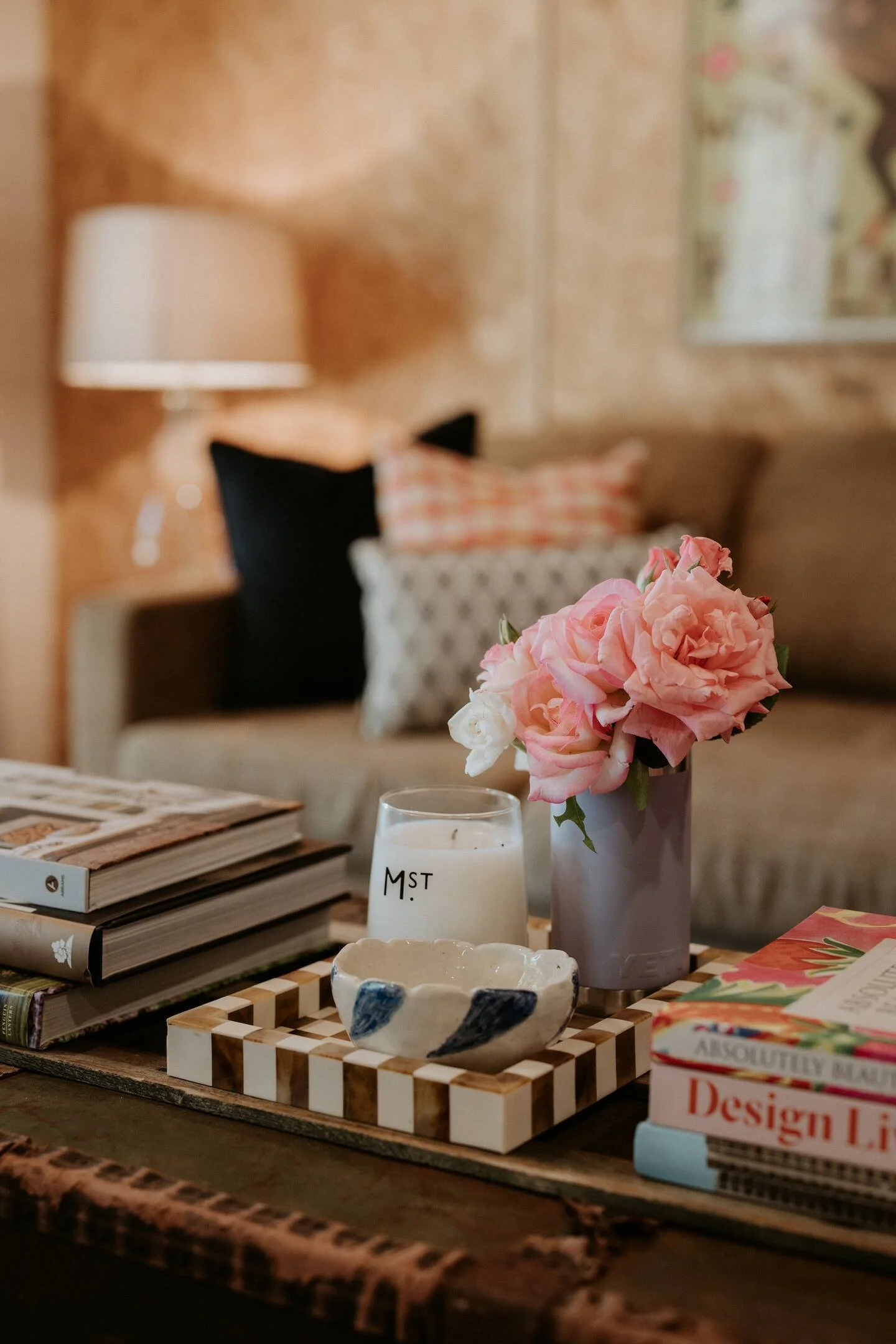 Close-up of a table with books, a ceramic bowl, a candle, and a vase with pink flowers, in a cozy living room setting.