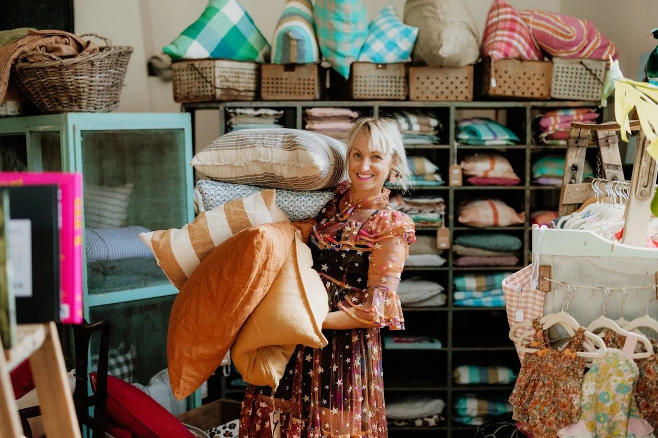 Woman shopping for pillows at a home decor store, holding several colorful pillows, with shelves of textiles and home accessories in the background.