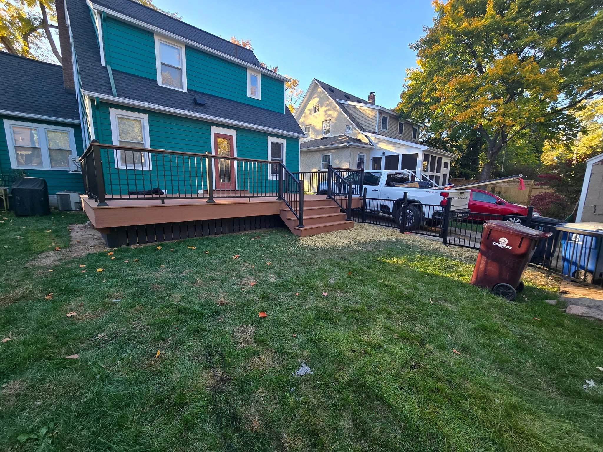 Backyard with a newly built wooden deck with stairs, a blue house, parked pickup truck, red car, and large trees in the background.