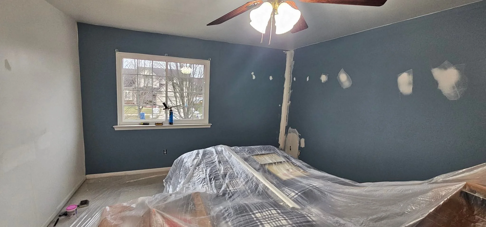 A bedroom under renovation with blue accent wall, window showing outdoor scene, and marking patches on the wall. Bed covered with plastic, ceiling fan with lights turned on.