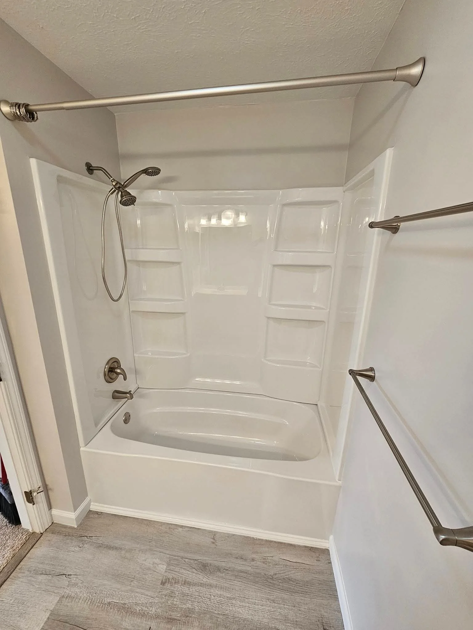 A white bathtub with a showerhead, drain, and silver faucets, surrounded by a white shower wall with built-in shelves, and two silver towel bars on the adjacent wall, with a ceiling-mounted shower curtain rod above.
