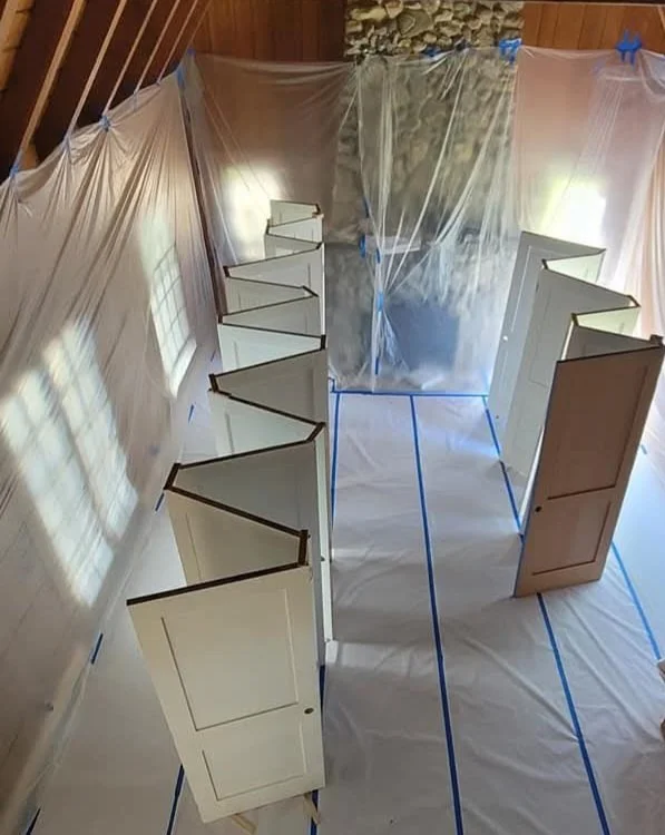 Construction or painting setup with white doors arranged on a floor covered with a plastic sheet, plastic sheeting on the walls, and blue painter's tape along the seams.