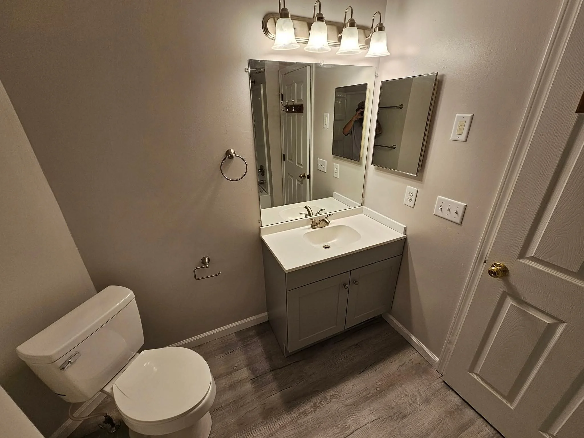 Small bathroom with a toilet, a sink with a cabinet, a large mirror, a small cabinet and several electrical outlets on the wall, a light fixture with five bulbs, and a door.