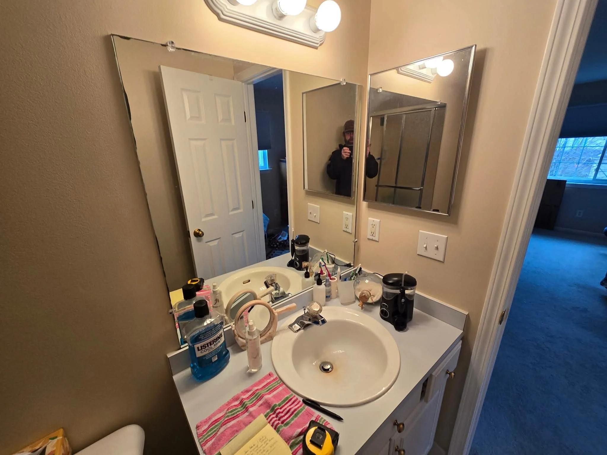 Bathroom bathroom sink with clutter, including hand sanitizer, toothbrushes, a hairbrush, a tape measure, and a water bottle; mirror above the sink reflects a person taking a photo; open door with closet behind; part of bedroom visible through doorwa