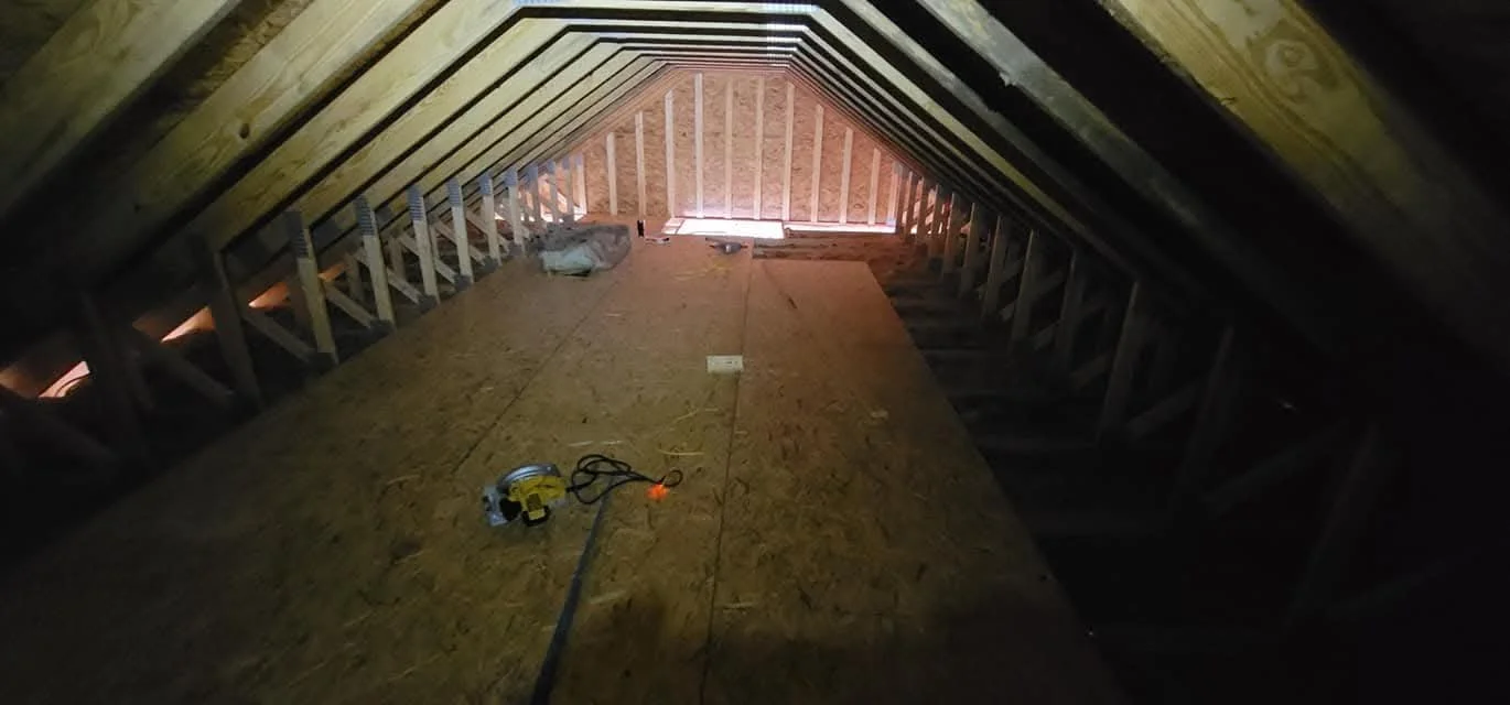 Attic under construction with exposed wooden framing and plywood flooring, tools and materials scattered inside.
