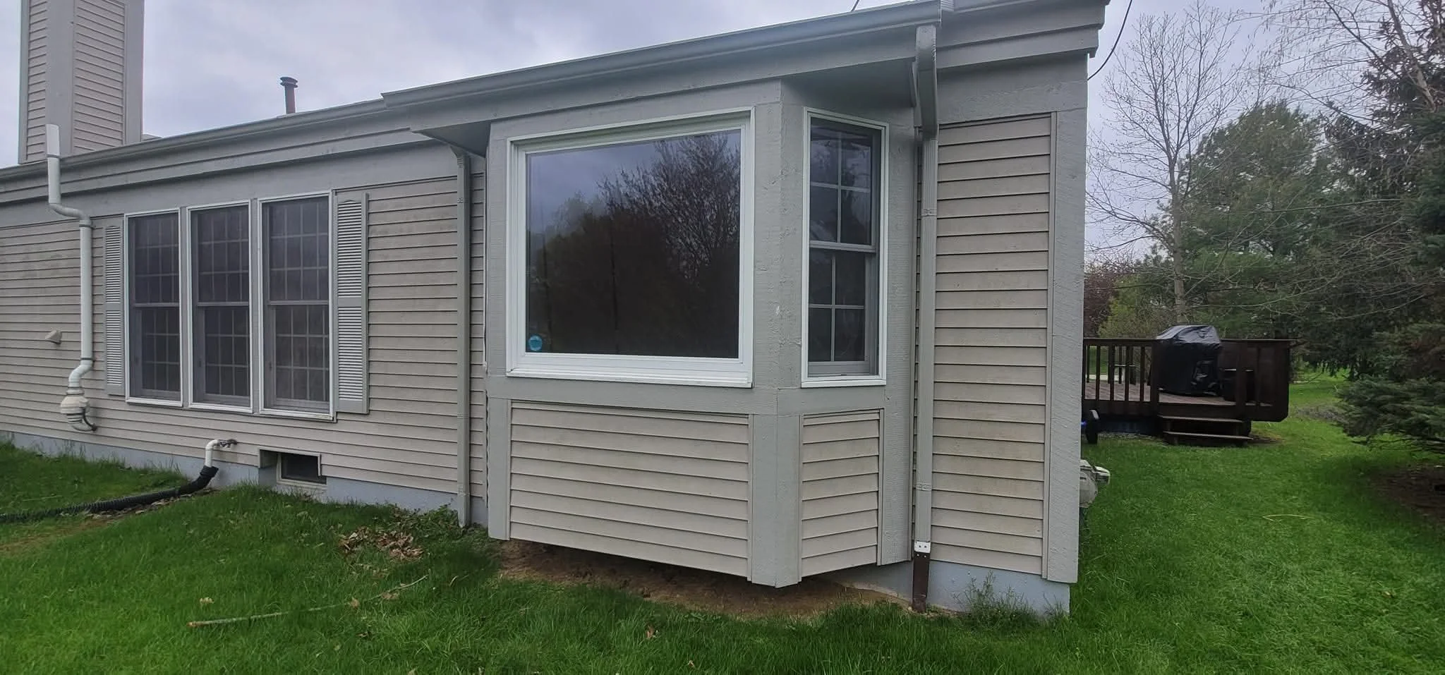 The back of a house with beige siding, a large window, a small window, a wooden deck with a grill, and a green grassy yard.