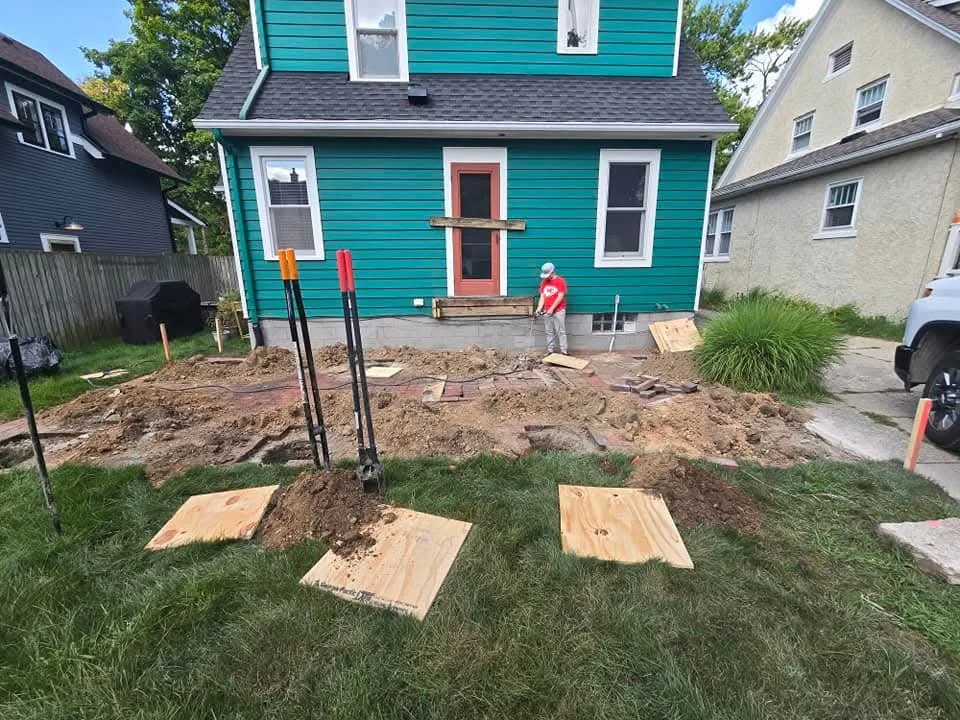 A person working on construction or landscaping in front of a teal house, with tools and plywood boards on the ground, dirt excavation, and a partially built porch.