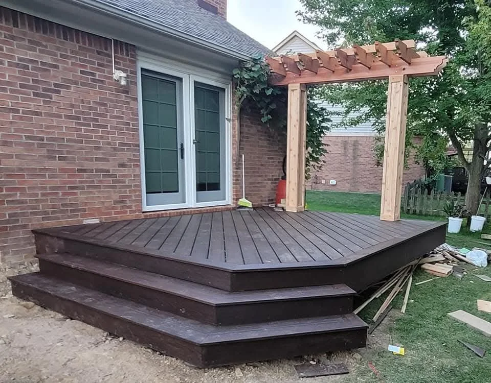 A newly constructed wooden deck attached to a brick house with a sliding glass door, featuring a pergola frame on one side in a backyard.
