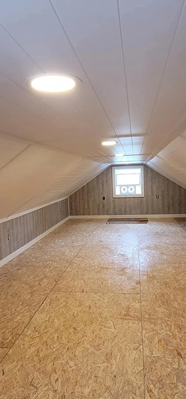 Empty room with slanted ceiling, wood-paneled walls, a small window, and unfinished plywood flooring.