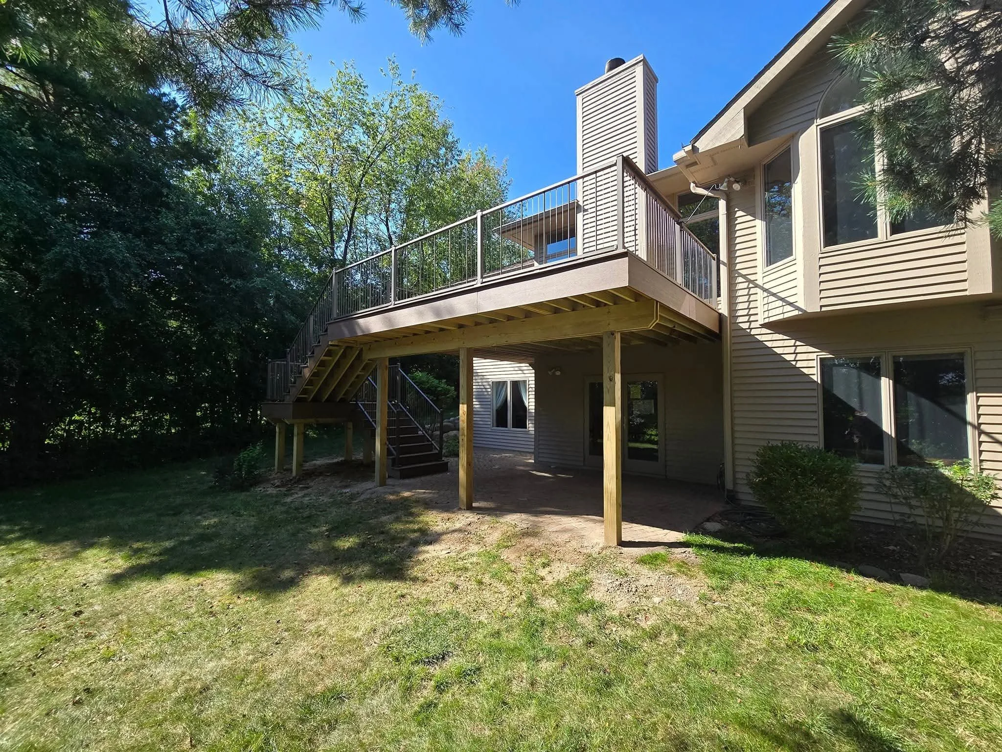 Newly built wooden deck with stairs and a railing attached to the back of a beige house with multiple windows, surrounded by green trees and grass on a sunny day.