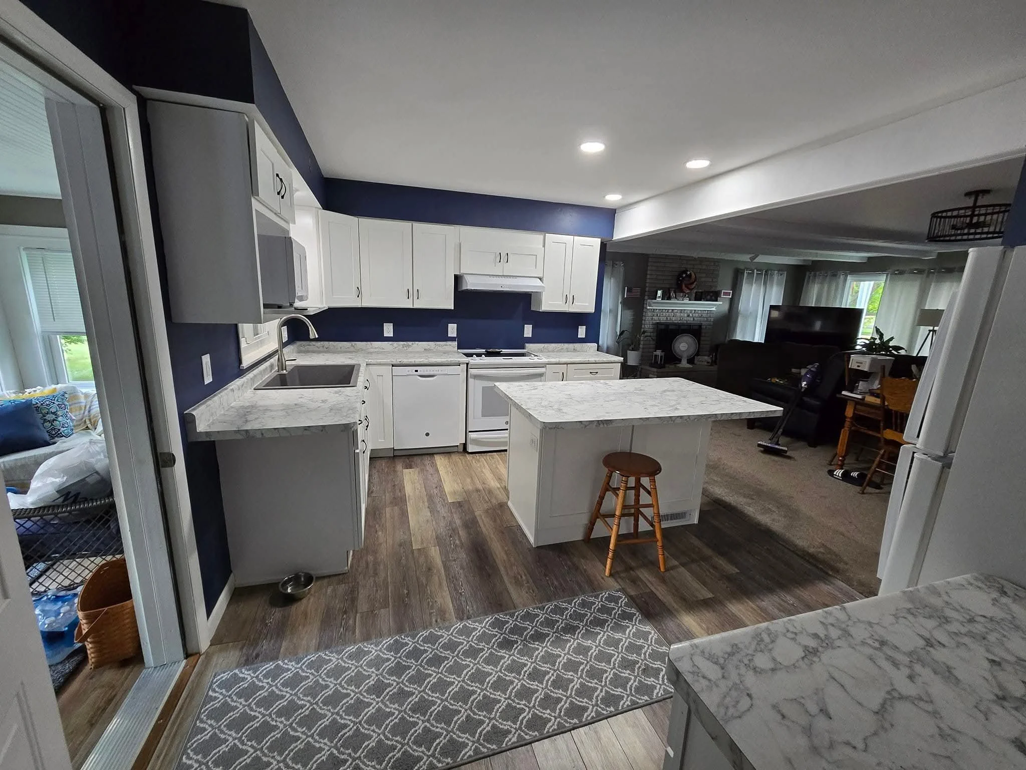Modern kitchen with white cabinets, marble countertops, a small island with a stool, and a view into a living room with a fireplace and a TV.