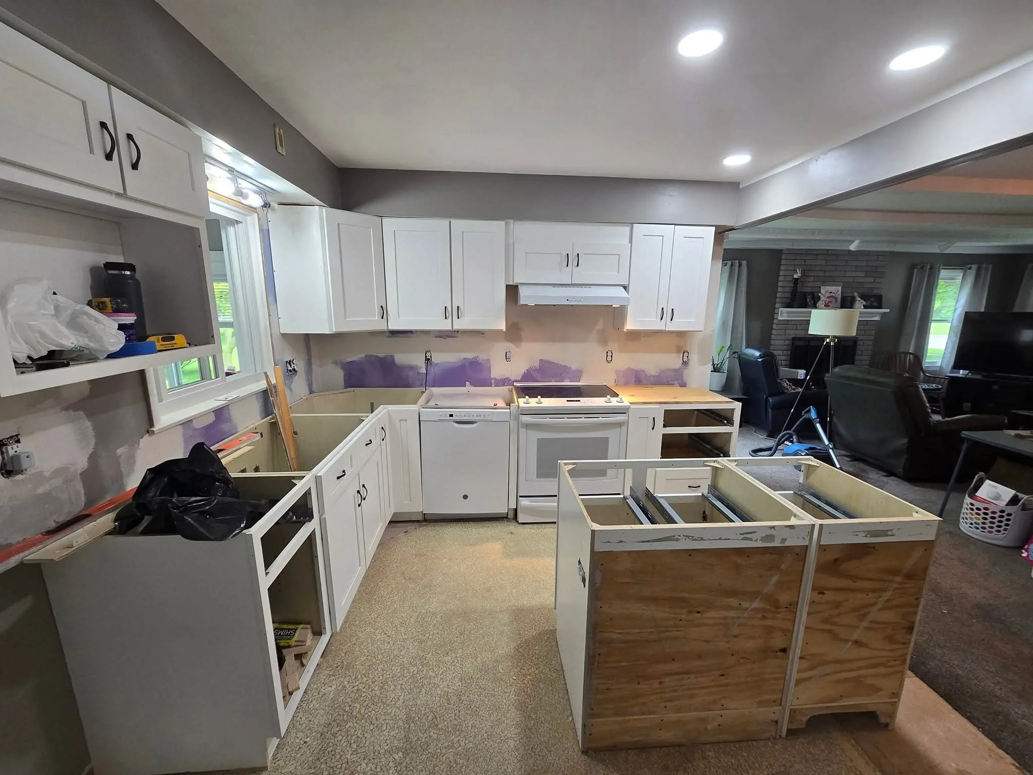 Kitchen under renovation with partially installed white cabinets, purple wall patching, and construction materials. Open living room with furniture and TV visible in background.