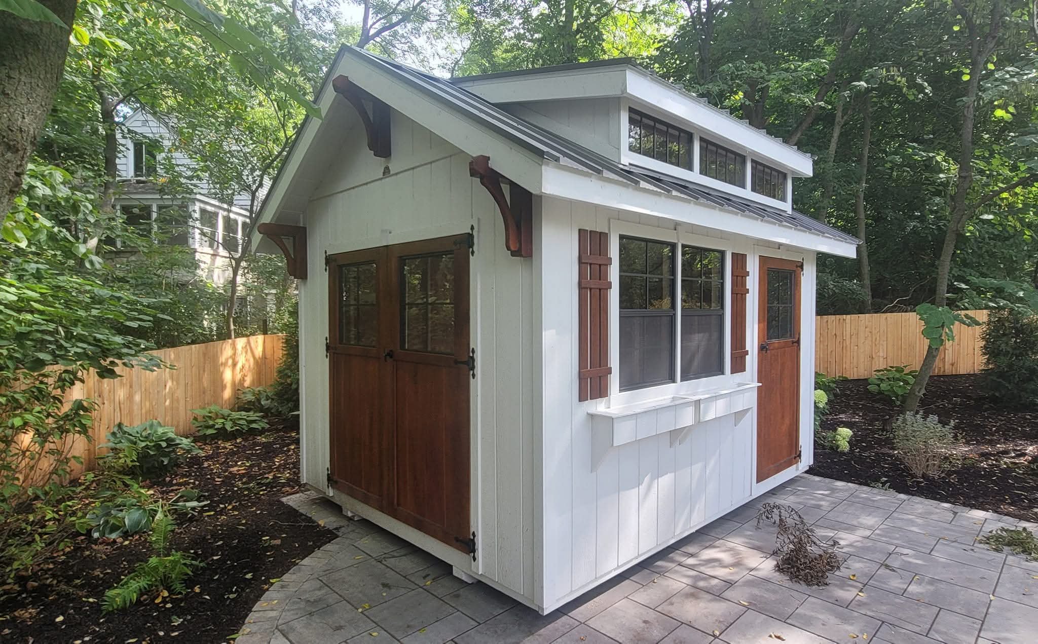 Small white outdoor shed with wooden doors, brick window shutters, and a sloped roof, situated on a paved area in a garden with trees and a wooden fence in the background.