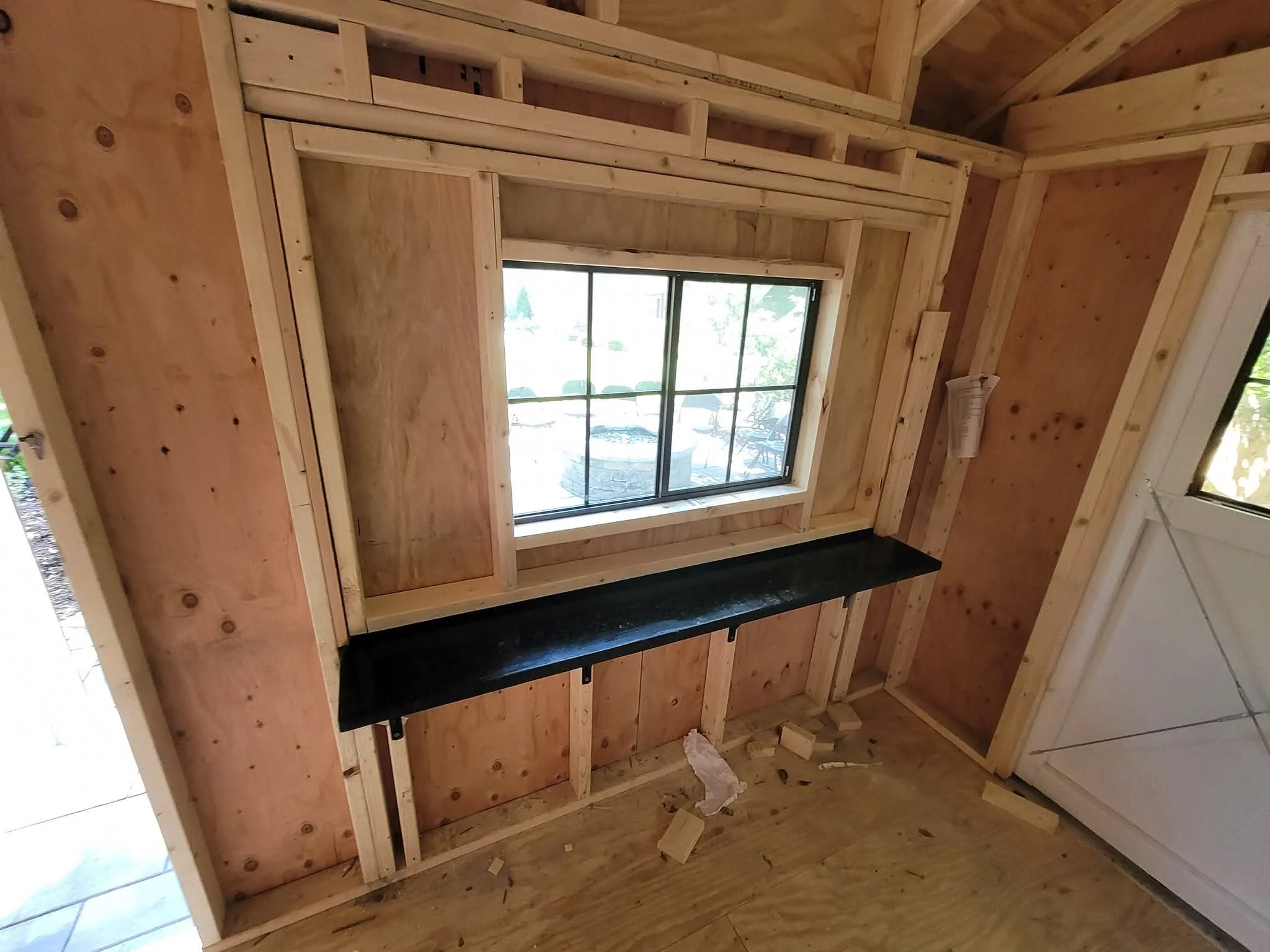 Interior view of a partially built room with wooden framing, a window, and a black countertop or shelf installed beneath the window.