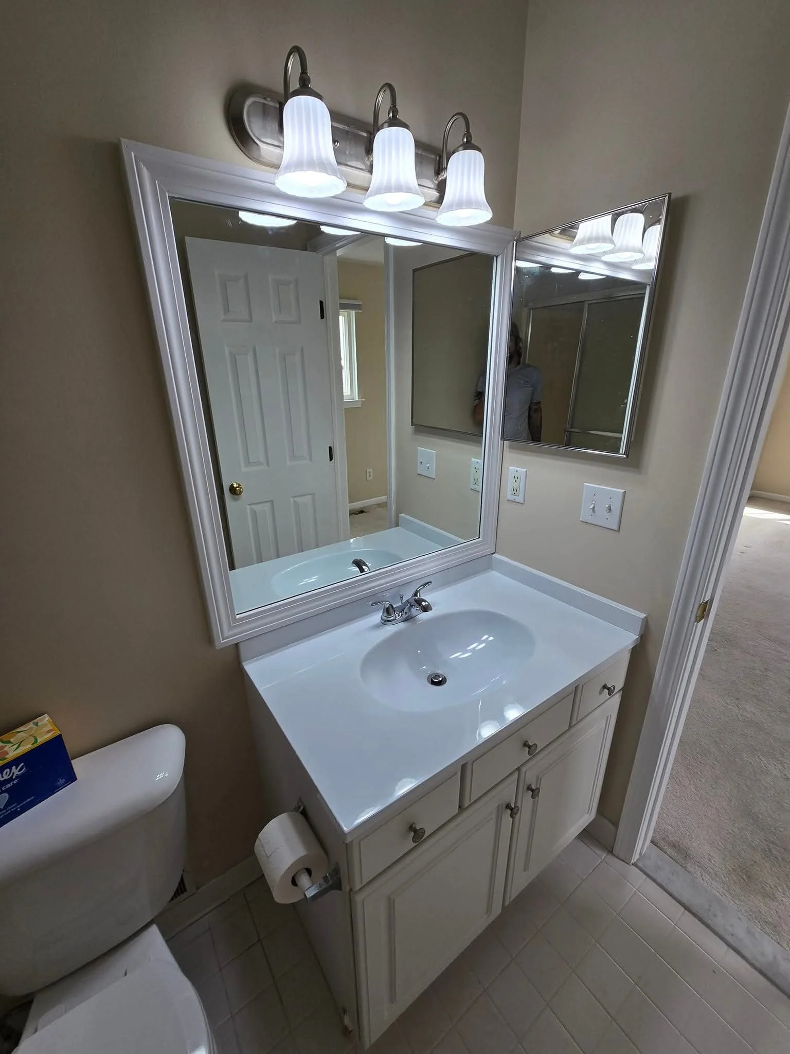 Bathroom vanity with a white countertop, oval sink, and a large framed mirror above. The light fixture has four white shades. Part of the toilet and bathroom door are also visible.