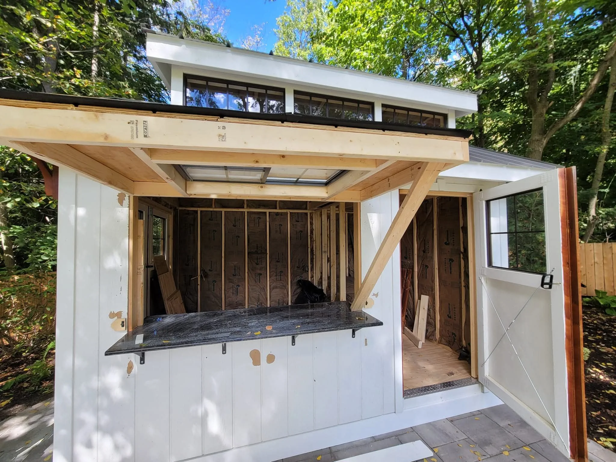 A small house under construction with an open door and large window, surrounded by trees, with construction materials and exposed unfinished interior visible.