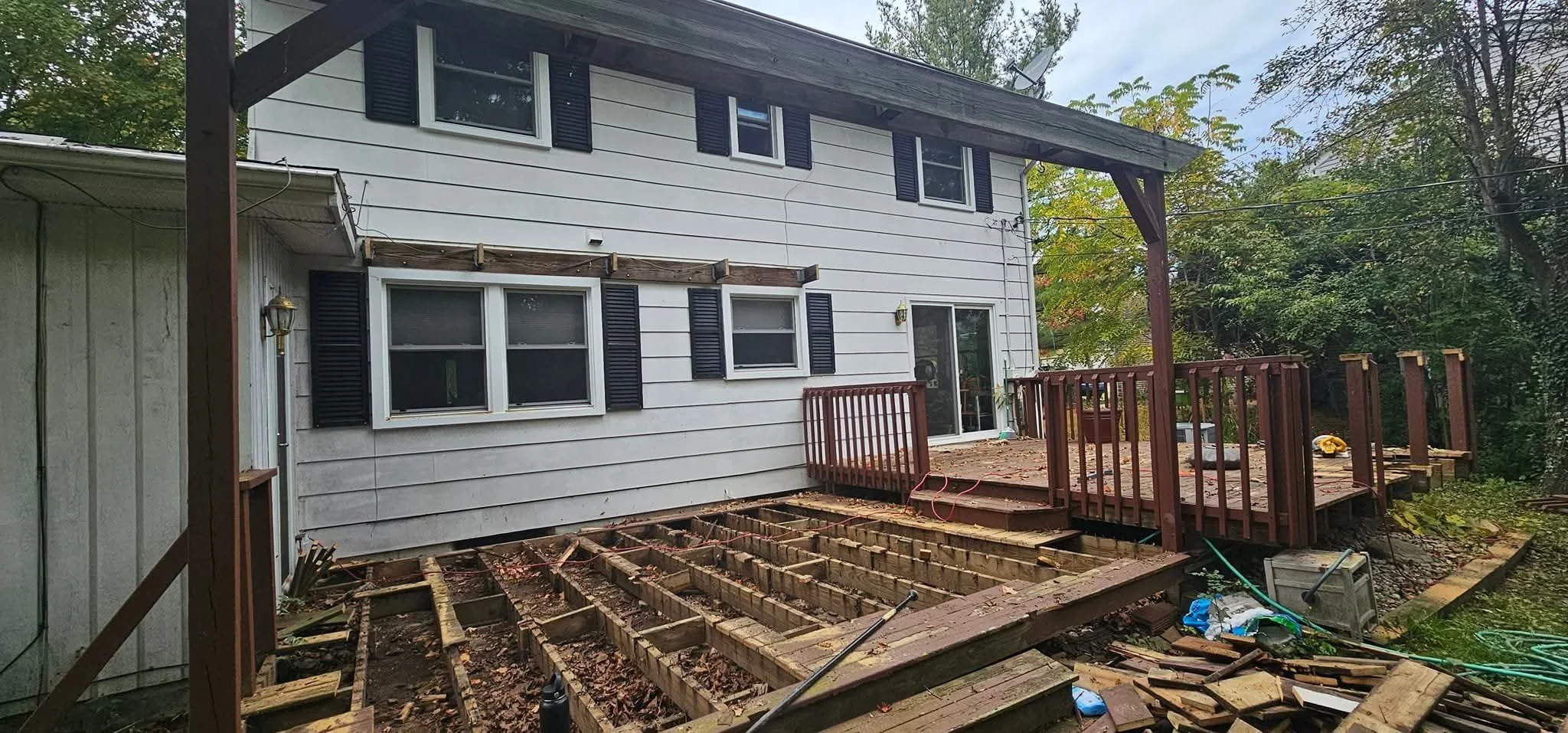 Backyard deck construction in progress with a partially built wooden deck and stairs, attached to a two-story house with white siding and black window shutters, surrounded by trees.