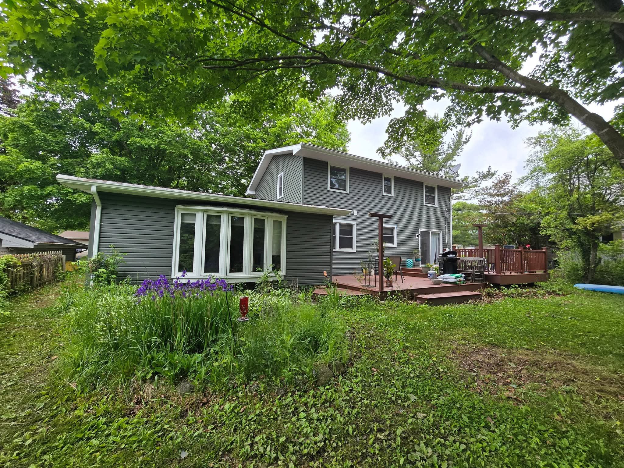Suburban backyard with green grass, flowering plants, mature trees, and a two-story home with new vinyl siding and composite deck