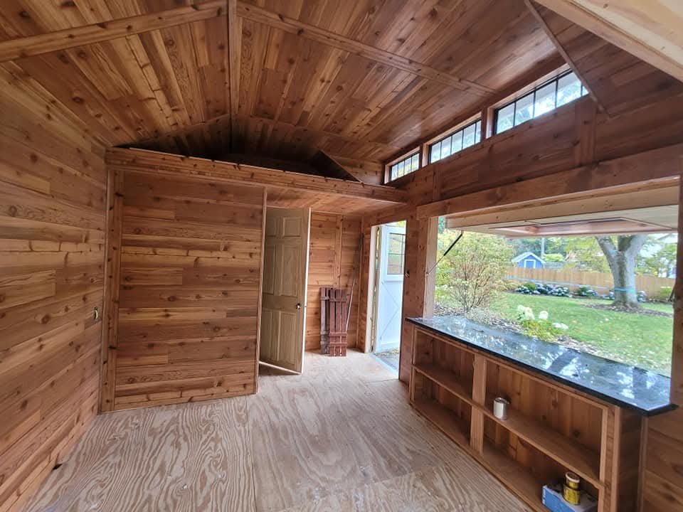 A small wooden room with unfinished walls, ceiling, and floor. There are windows near the ceiling, a door leading outside, and a black countertop with open shelving underneath. Outside, a backyard with trees and a house is visible.