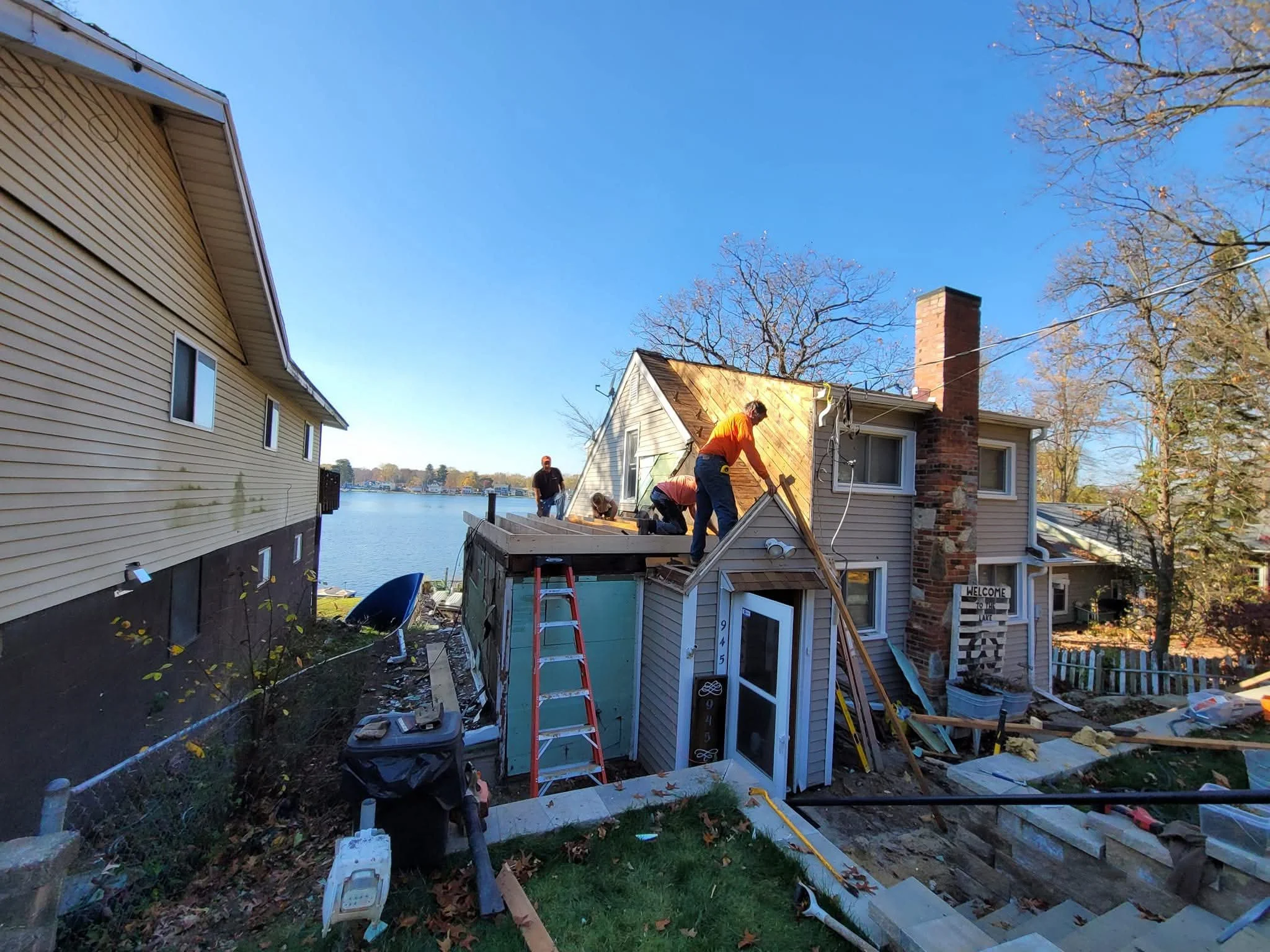 People working on extending the roof of a house by a lake, with a ladder and construction materials around.
