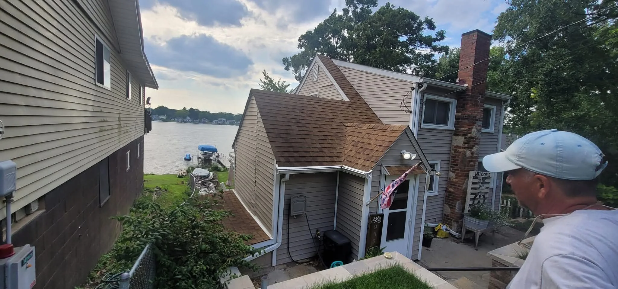 A man in a white cap and white shirt standing on a backyard patio overlooking a waterway with boats and houses. There is a house with a brick chimney nearby, and a small outbuilding with a brown roof. The scene is partly cloudy with some sunlight.