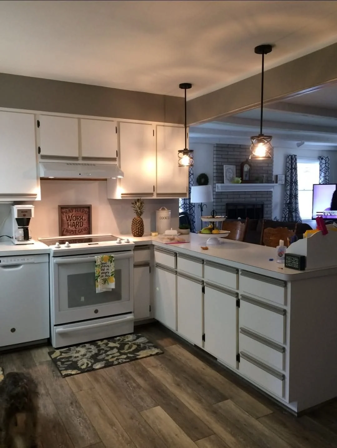 Kitchen with white cabinets, stove, pineapple decor, pendant lights, and a view of a living room with a brick fireplace.