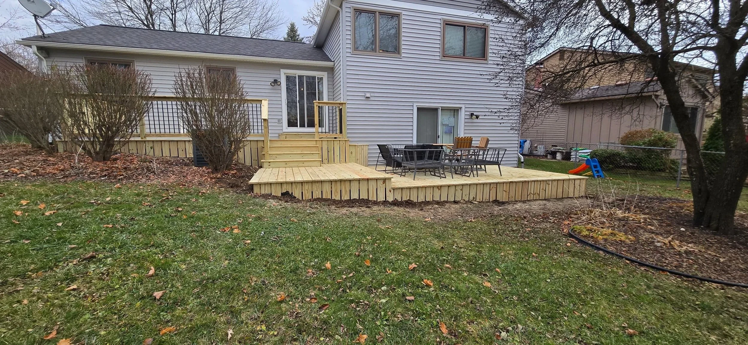 Backyard with a newly built wooden deck, patio furniture, and a children's slide, surrounded by trees and neighboring houses.