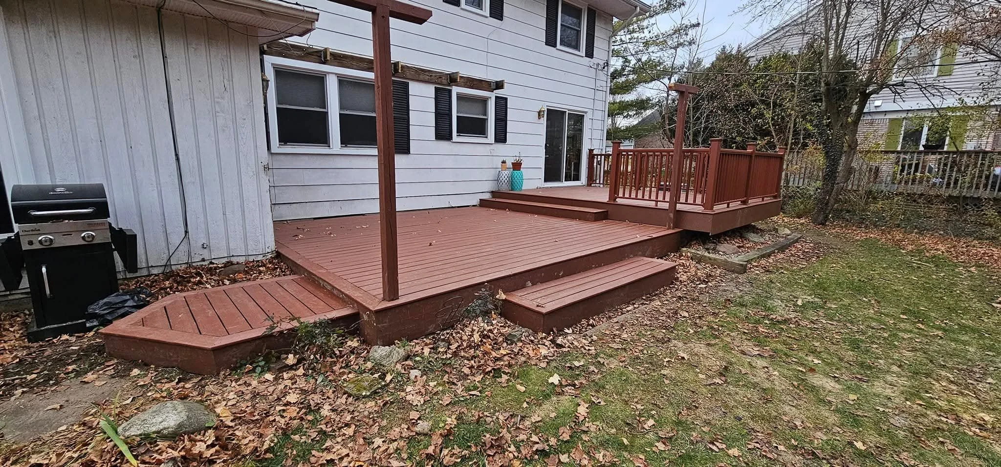 Backyard with a newly built wooden deck with steps, a small deck extension, and a grill on the left side near the house, with leaves scattered on the ground.