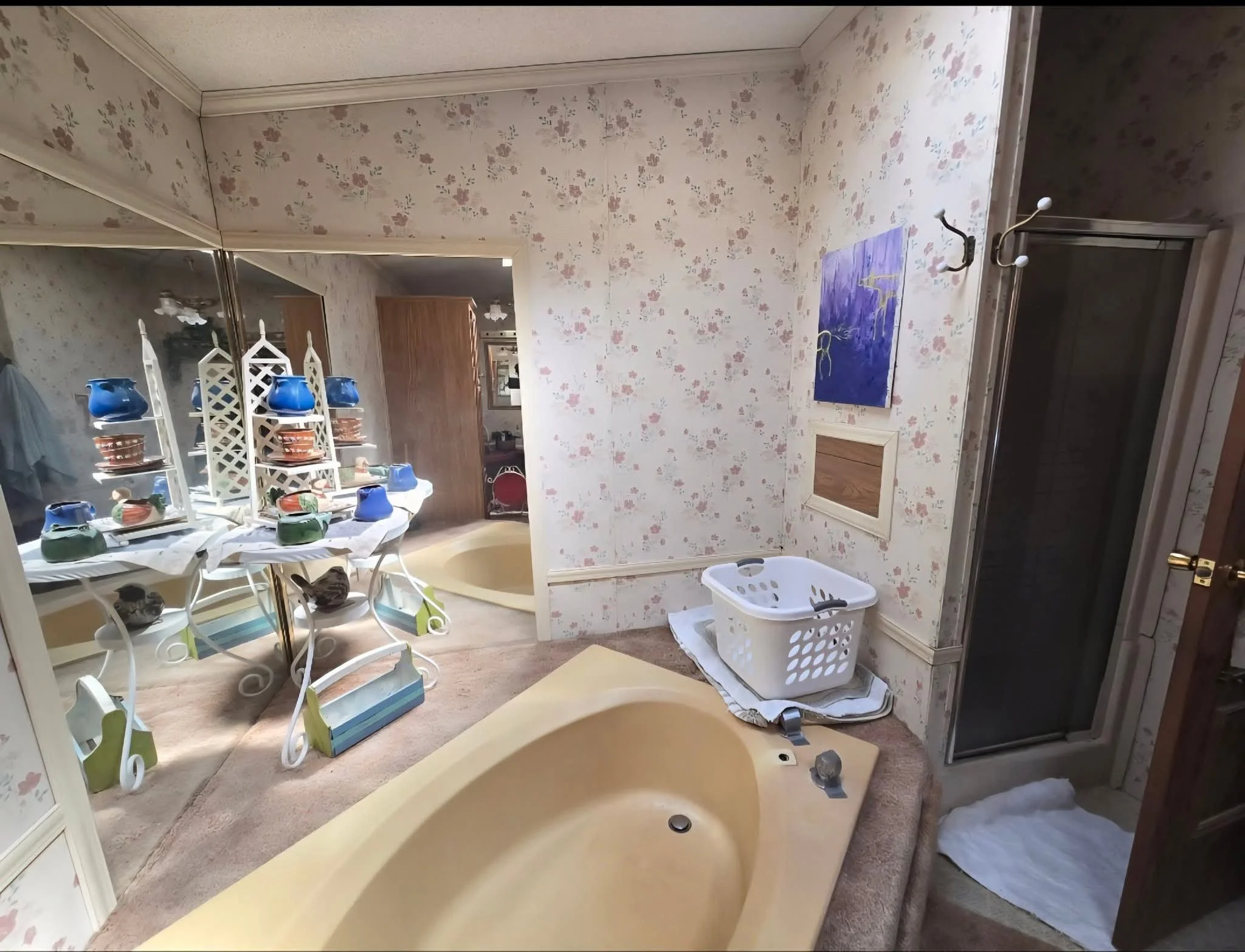 A bathroom with floral wallpaper, featuring a yellow bathtub, a shower enclosure, and a laundry basket. There is a mirror on the left reflecting a table with blue and orange decorative items and a room with chairs in the background.