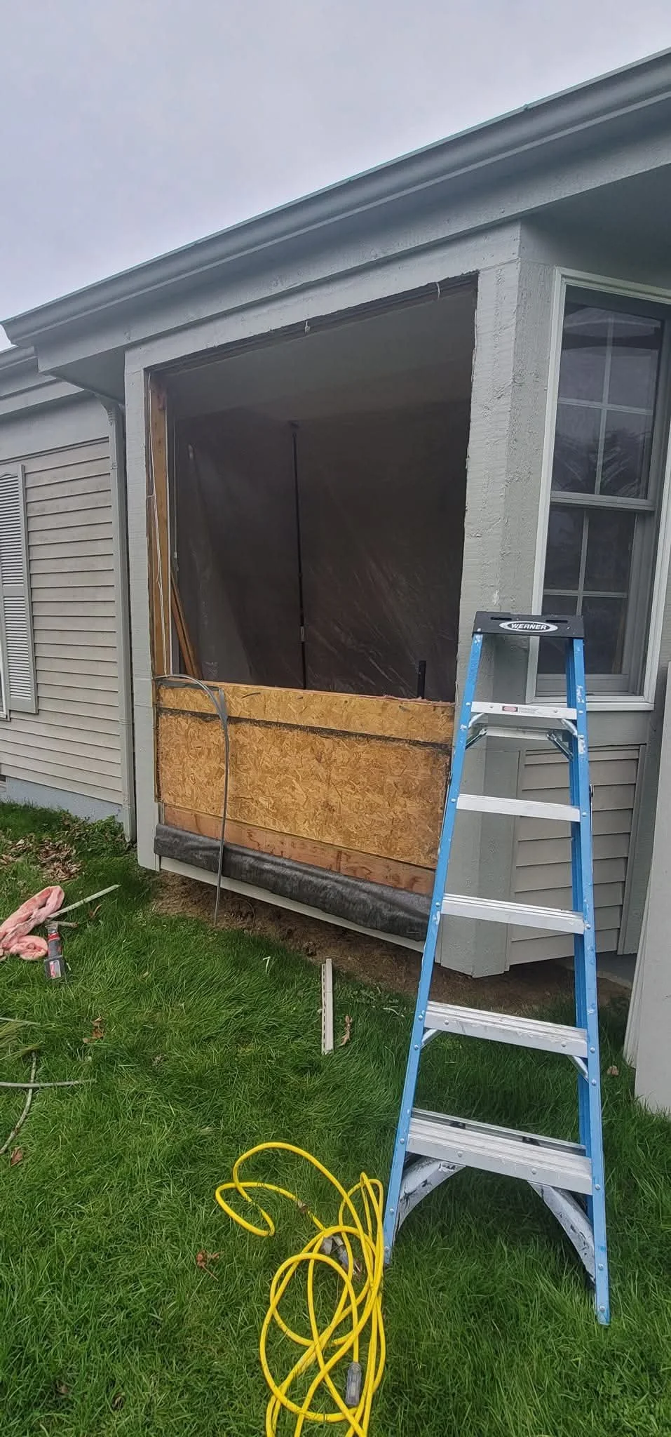 A house under renovation with a screened porch framing being installed, a blue ladder leaning against the house, and tools and yellow extension cord on the grass.