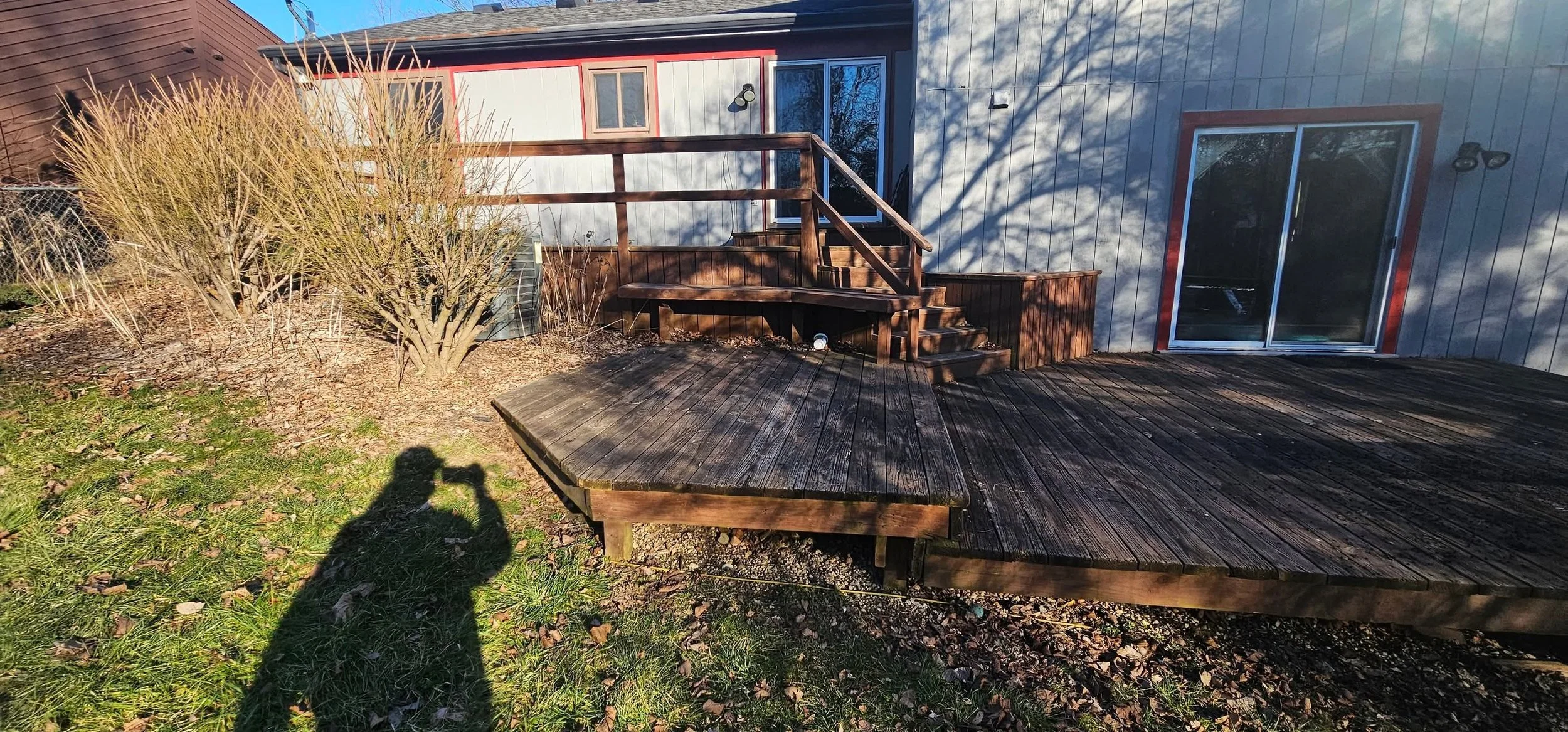A backyard deck with a wooden surface and railing, next to a house with sliding glass doors and a small window, and a bush on the left side. The house has vertical siding and the photo shows shadows on the ground and the house.