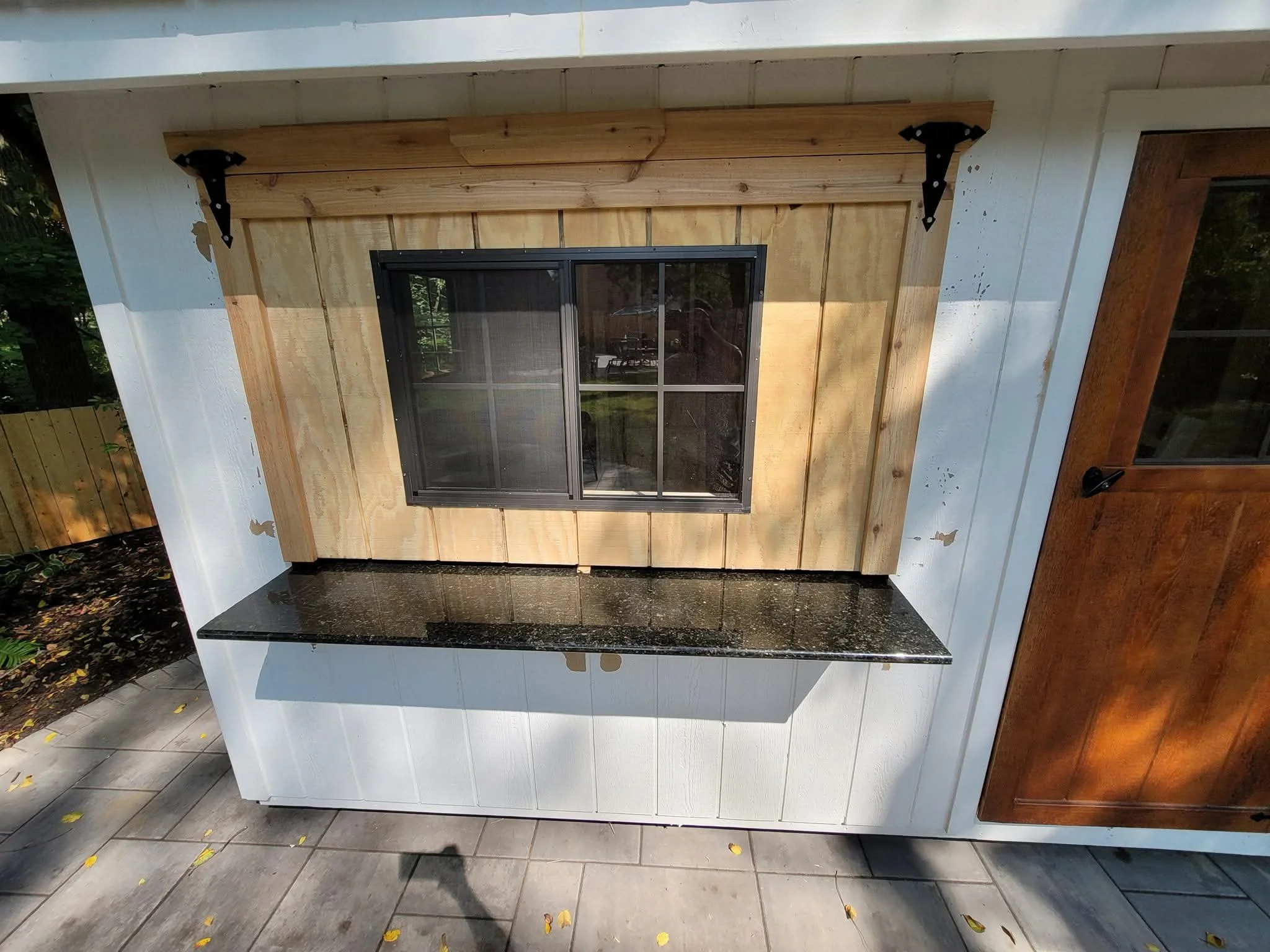 Exterior of a small building with a window, a granite countertop below the window, and a wooden door on the right. The siding is white, and there are black metal brackets holding a wooden trim above the window.