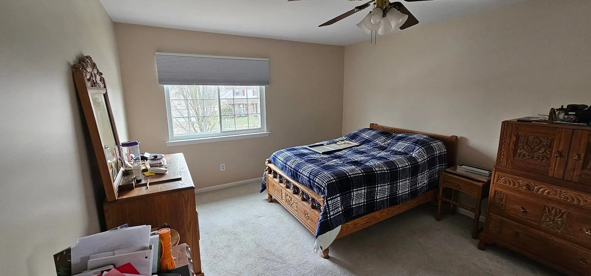 Bedroom with a wooden bed frame, plaid bedding, a dresser with mirror, a side table, and a window with a view of trees and houses outside. Freshly painted beige
