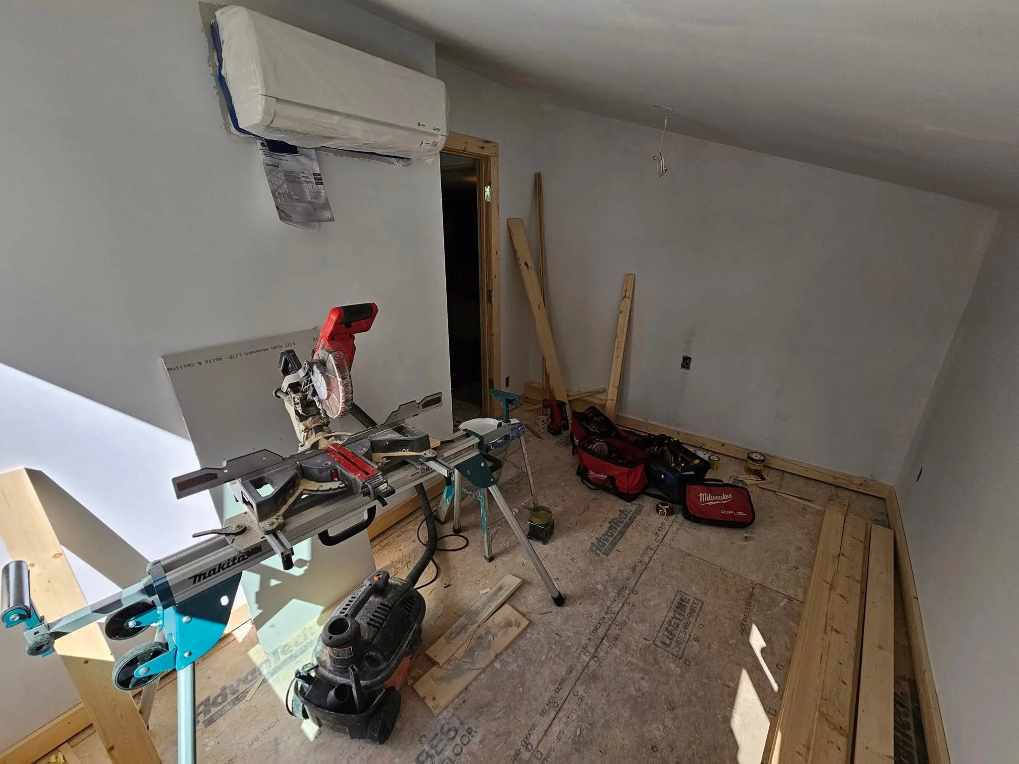 A room under construction with a miter saw on a stand, tool bags, lumber, and various tools on the floor. The walls are partially finished with drywall, and there's an air conditioning unit mounted on one wall.