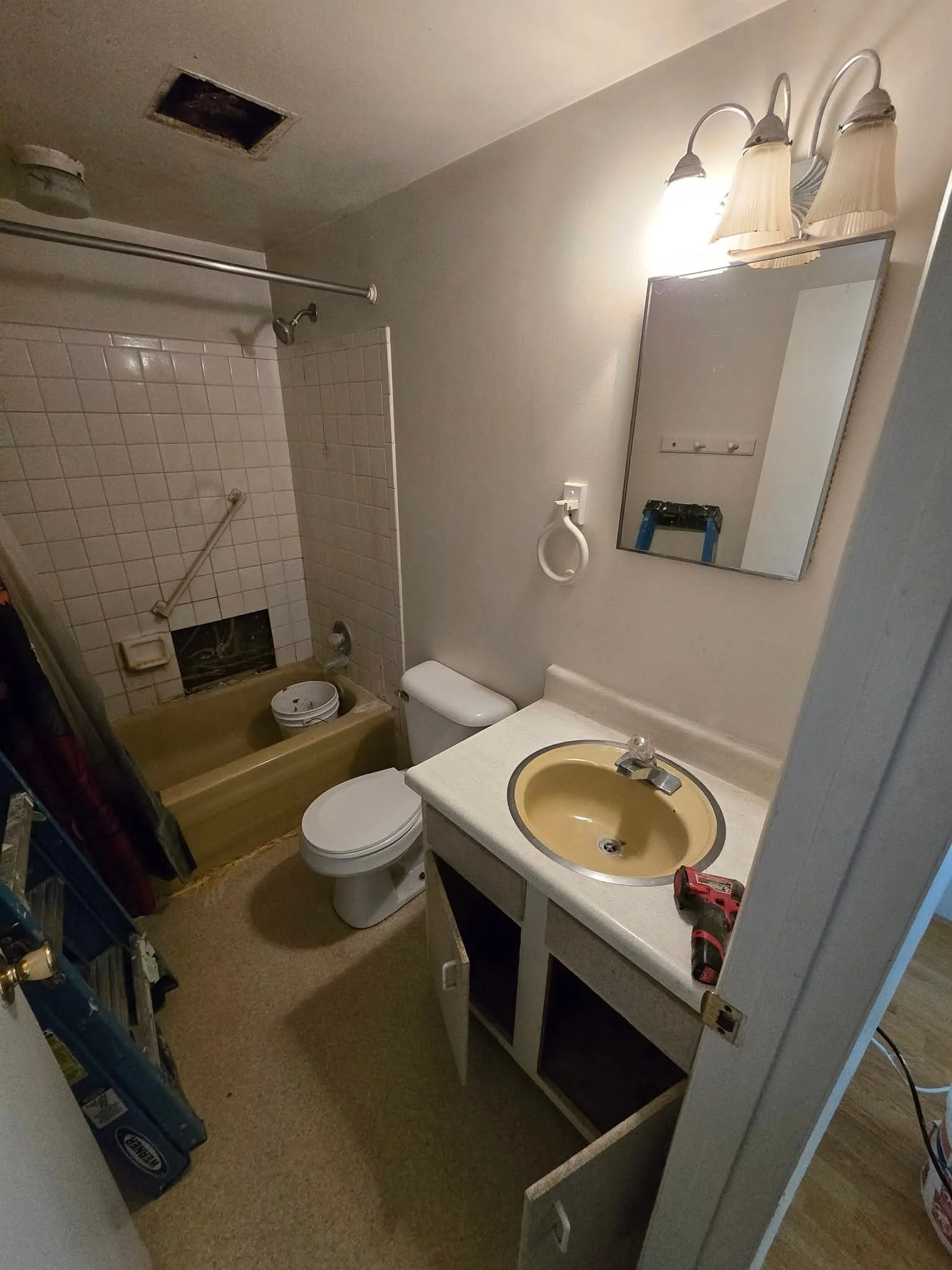 Small bathroom with a bathtub, toilet, and vanity with a yellow sink, mirror, and light fixture. The vanity door is open, revealing the inside. A power drill is on the counter. The ceiling has an open vent or access panel, and there is a towel ring n