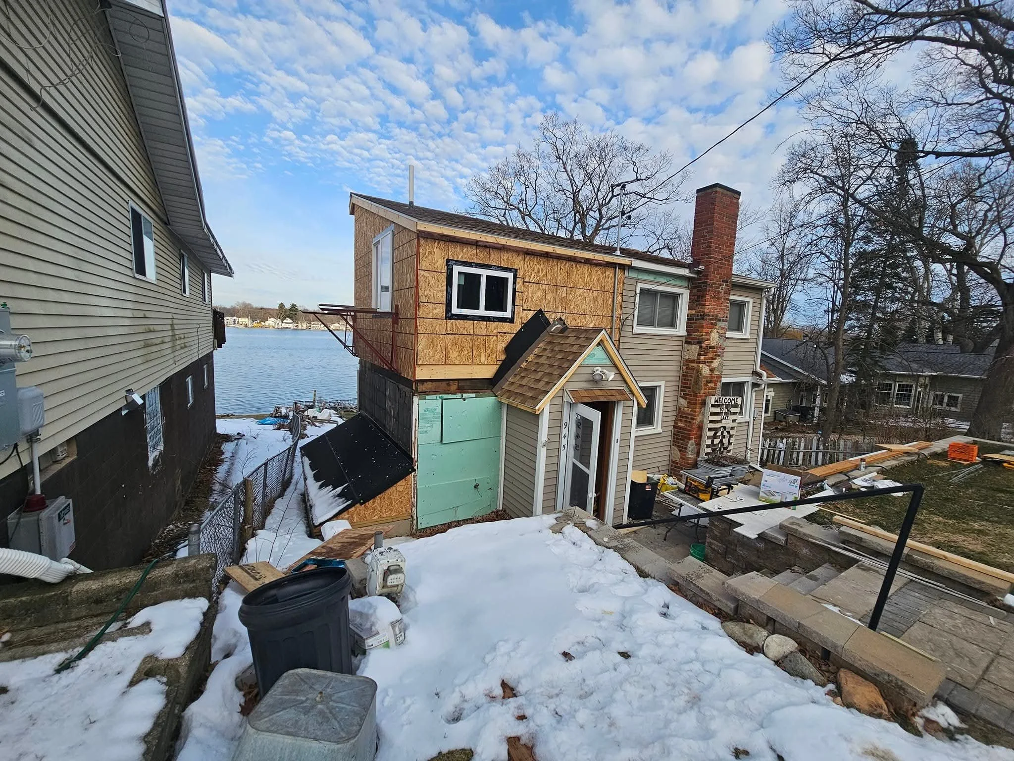 A house under renovation with a partially built second floor, a brick chimney, and snow on the ground near a lake