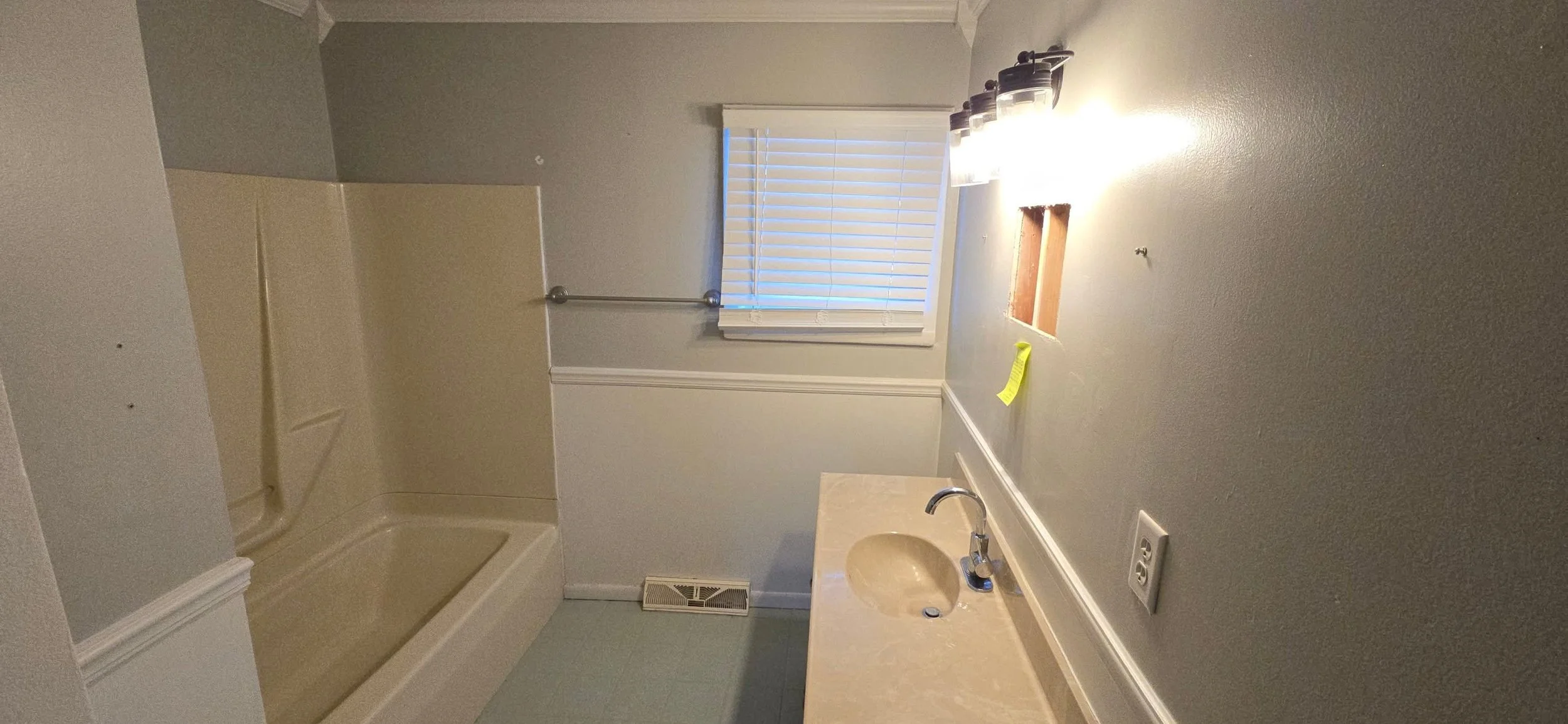 Empty bathroom with a beige bathtub and shower in the corner, a small window with blinds, a cream-colored countertop with a built-in sink, a wall-mounted light fixture above the sink, and a wall vent near the floor.