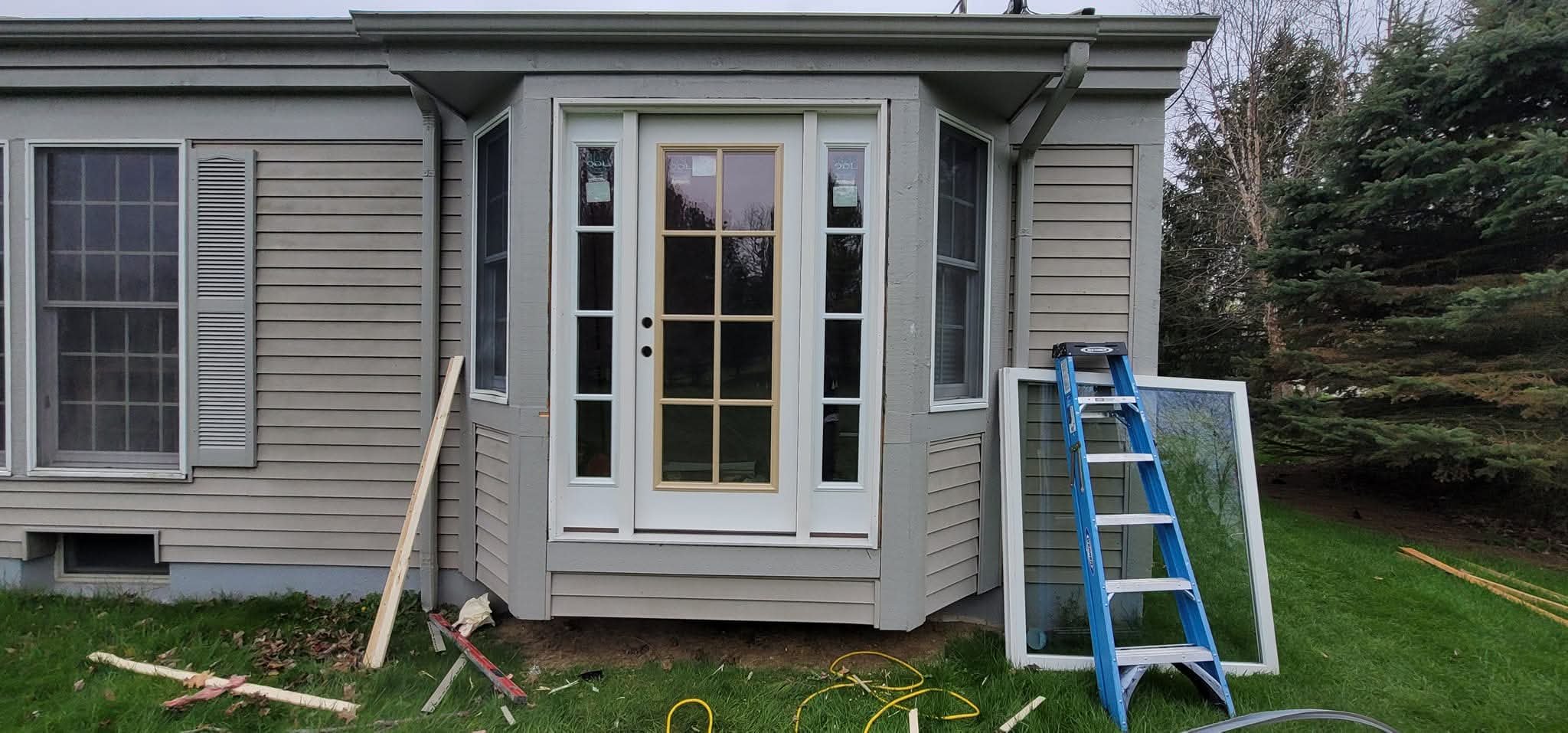 Construction of a backyard porch or sunroom with white framing and a door, a blue ladder leaning against the structure, a window frame on the ground, and construction tools and materials scattered on the grass.