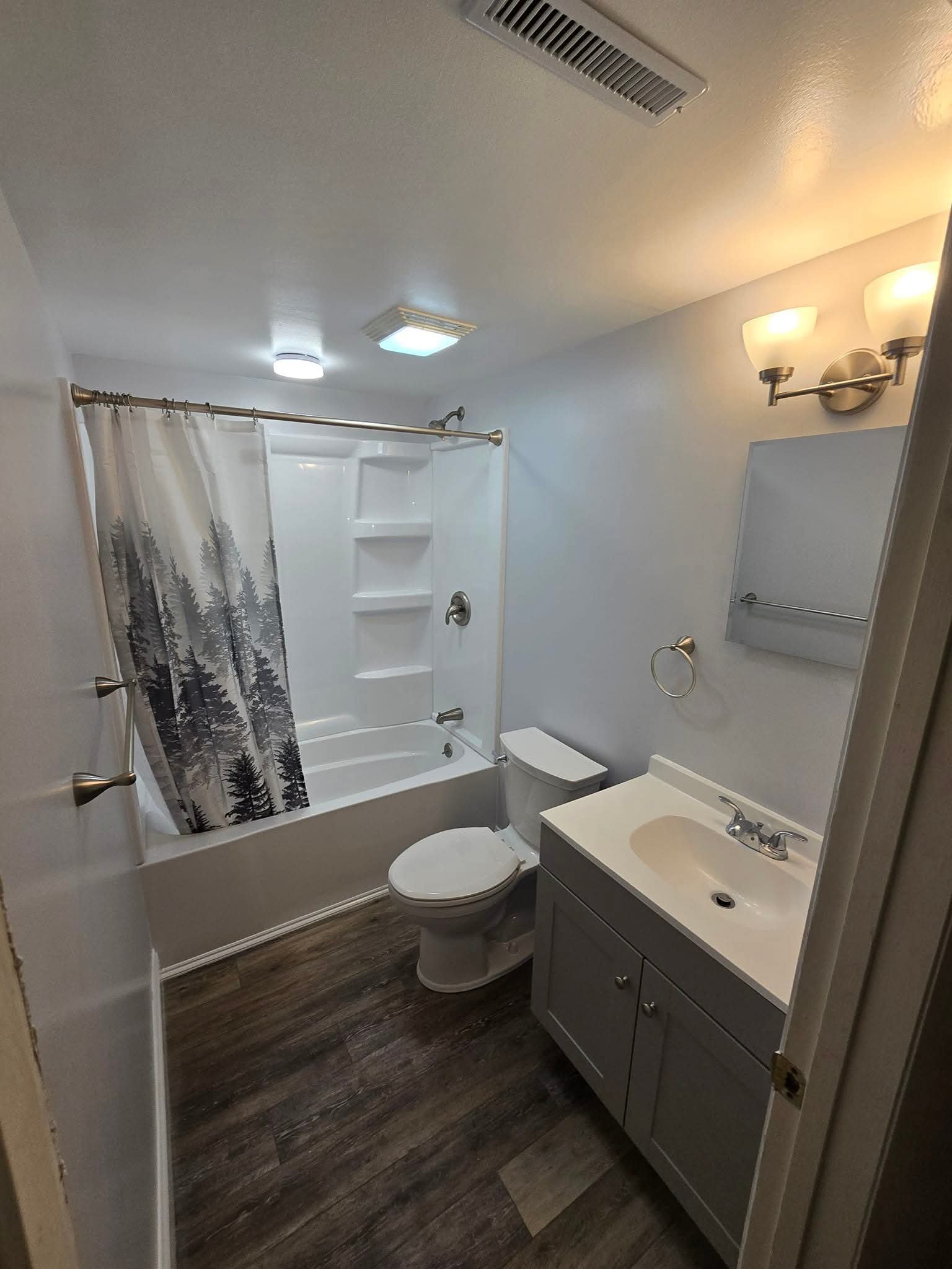 A small bathroom with a white bathtub and shower combination, a toilet, and a gray vanity with a white sink. There is a mirror above the vanity, a towel ring next to it, and a light fixture with two bulbs. The bathroom floor has wood-like tiles, and 
