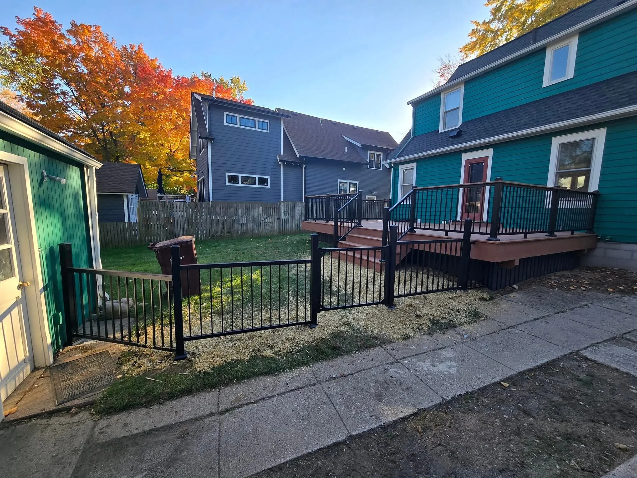 Backyard with a small fenced area, grass, a wooden deck with railing and stairs, and neighboring houses under a clear sky with autumn trees.