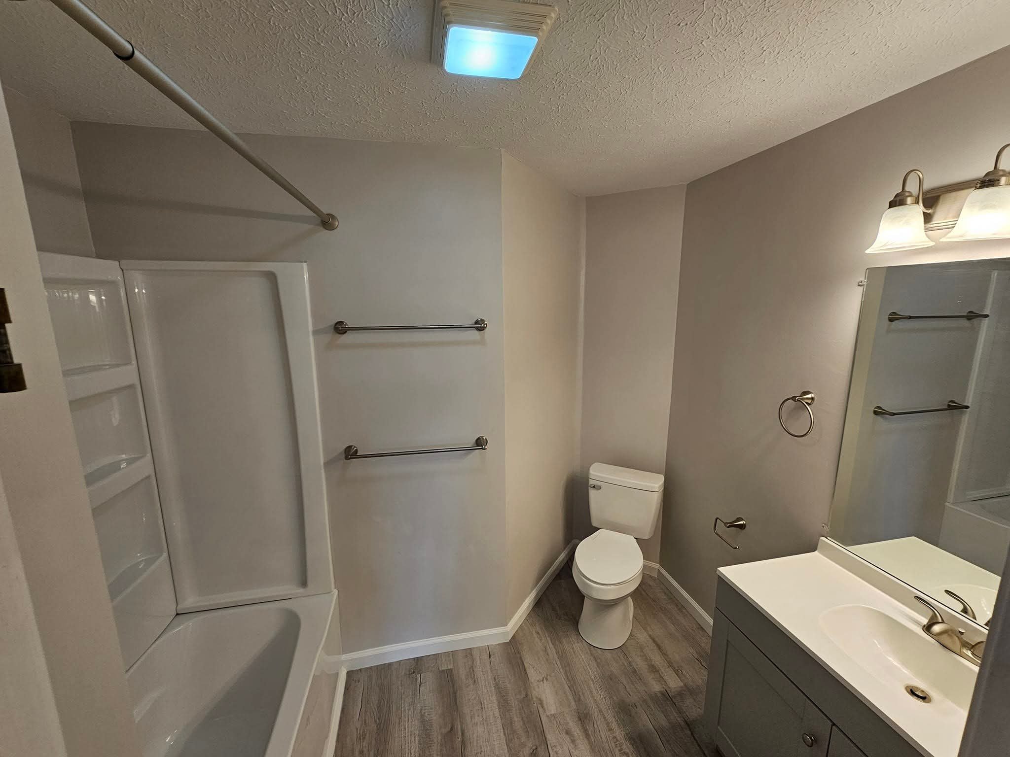 A small, clean bathroom with a white bathtub and shower, a toilet, a mirror with a light fixture above it, and a white vanity with a sink. The floor is wood-textured, and the walls are light-colored.