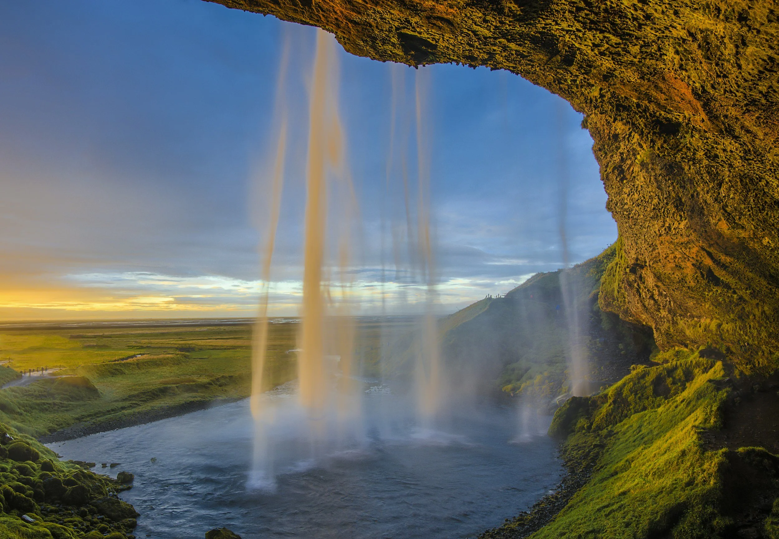 Waterfall cascading from a rocky overhang into a river below, with a green landscape and a sunset sky in the background.