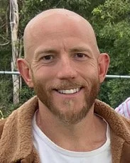 A man outdoors in a wooded area, smiling, wearing a dark hoodie.